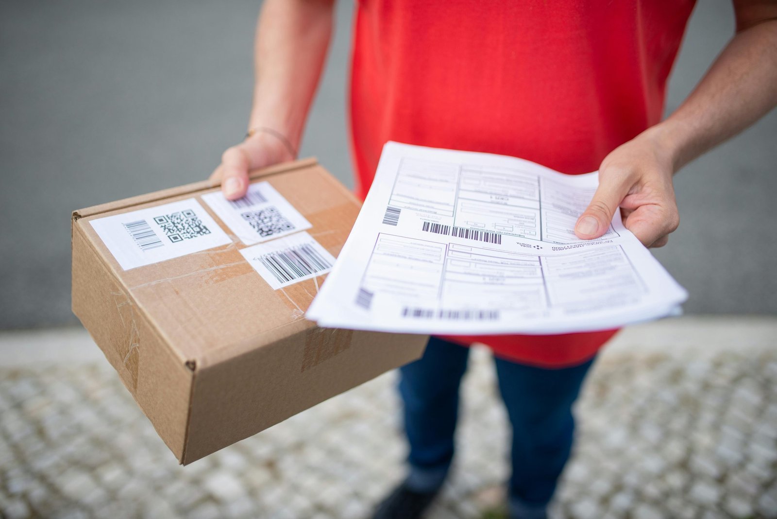 Close-up of person in red shirt holding a brown package and delivery documents outside.